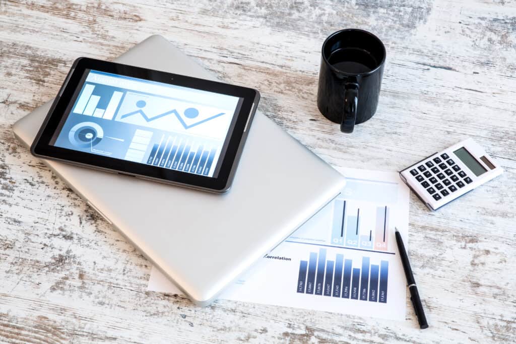 Tablet displaying financial graphs and charts placed on a laptop, alongside a calculator, pen, and coffee cup, symbolizing cost management and analysis.